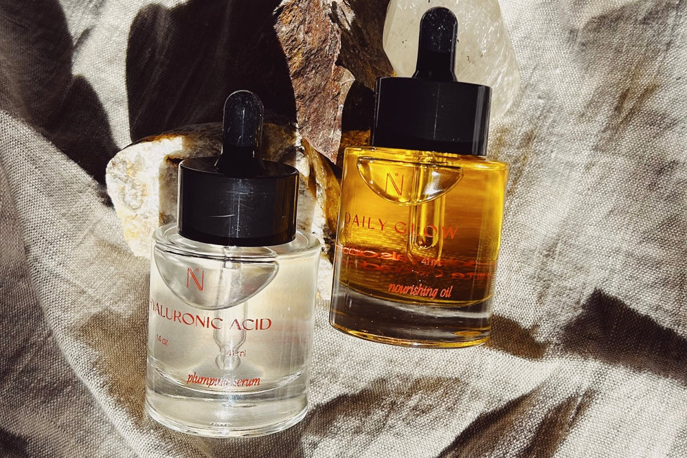 Two skincare bottles, one clear with a dropper labeled 'Niacinamide' and the other amber with a dropper labeled 'Nourishing Oil', on a textured fabric background.